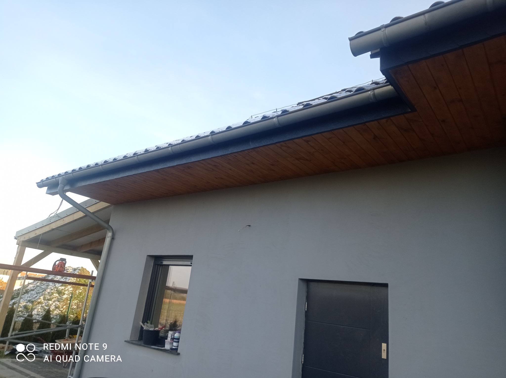 Szary dom jednorodzinny z widoczną drewnianą podbitką dachową, orynnowaniem i fragmentem elewacji w trakcie wykańczania, z rusztowaniem w tle.