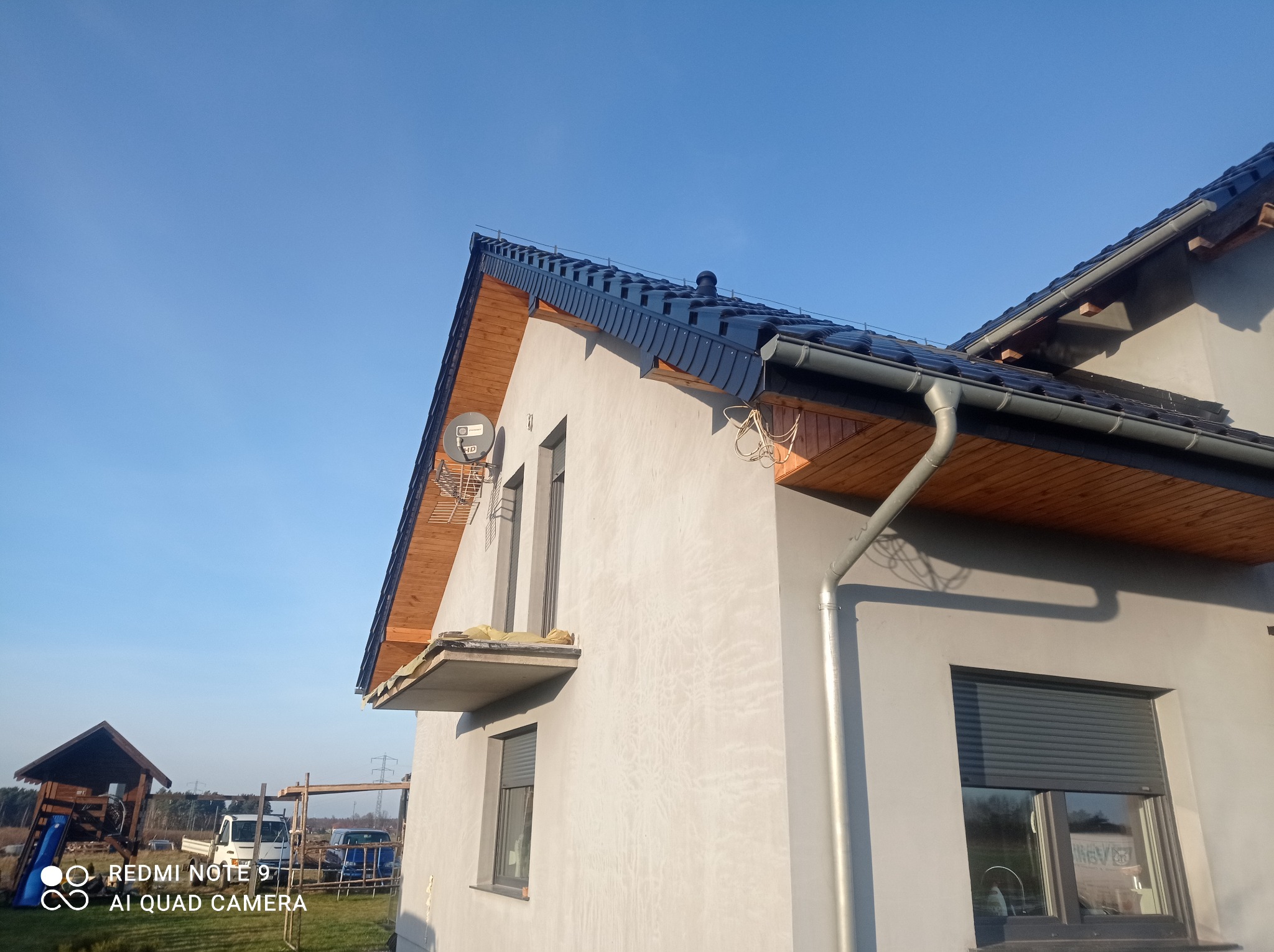 Nowy dom jednorodzinny z ciemną blachodachówką, drewnianą podbitką i szarą elewacją w trakcie wykańczania, widok z dołu na tle błękitnego nieba.