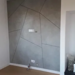 Ściana z szarą gładzią dekoracyjną z geometrycznym wzorem, widoczne gniazdka elektryczne, drewniana podłoga, czarne lampy sufitowe.