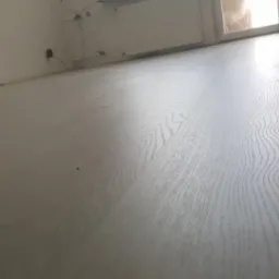 Jasnoszara podłoga z paneli laminowanych z wyraźnym wzorem drewna, ułożona w nowym mieszkaniu, widok z niskiego kąta, w tle widoczne okno i fragment ściany.