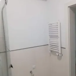 Jasna łazienka z kabiną prysznicową i grzejnikiem drabinkowym, wykończona płytkami ceramicznymi z szarym paskiem dekoracyjnym, widok z niskiego kąta.