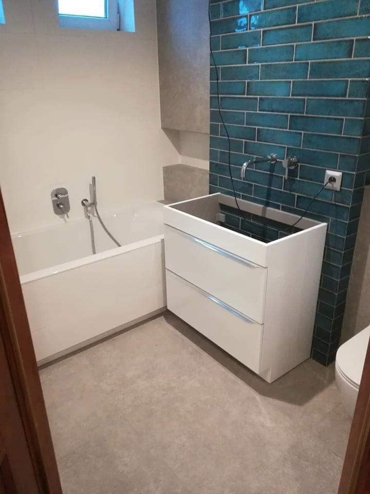 Niedokończona łazienka z turkusową ścianą z płytek, białą wanną i szafką pod umywalkę bez blatu, widoczne kable i gniazdko elektryczne.