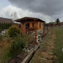 Wykop pod fundament ogrodzenia z drewnianymi słupkami w trakcie realizacji, widoczny sznurek wyznaczający linię, w tle drewniany domek letniskowy i pochmurne niebo.