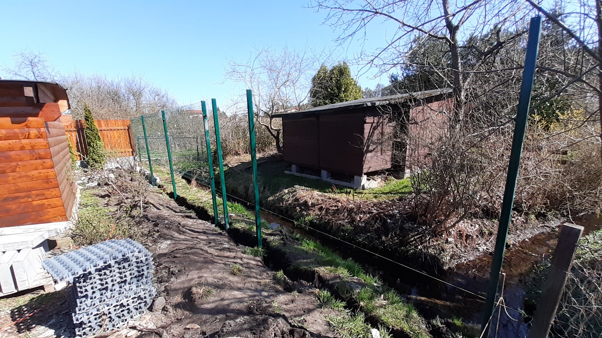 Wkop pod ogrodzenie z zielonymi słupkami wzdłuż strumyka, widoczny stos paneli ogrodzeniowych i drewniana zabudowa w tle.