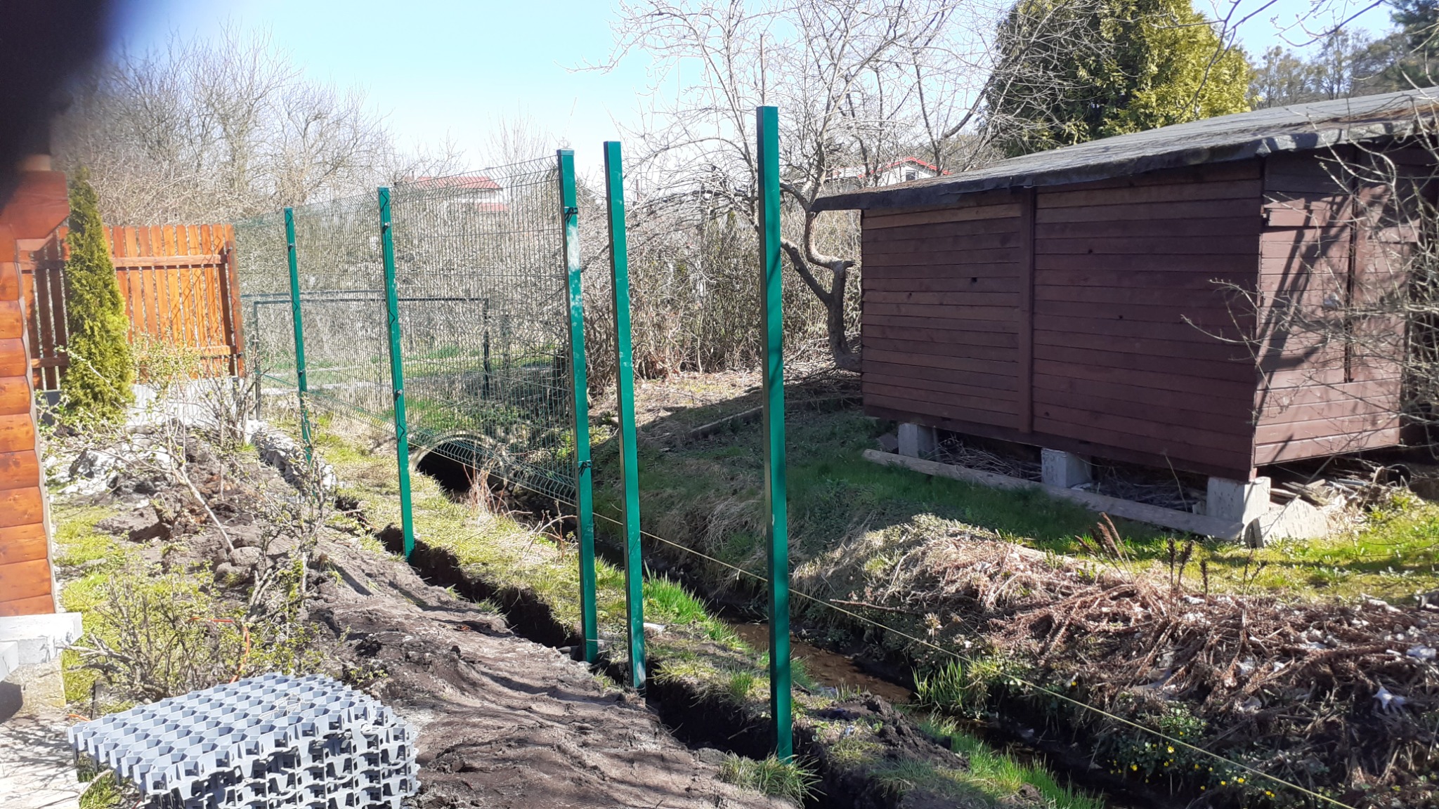 Wykopany rów pod siatką ogrodzeniową z widocznymi zielonymi słupkami, w tle domek narzędziowy i drzewa bez liści.
