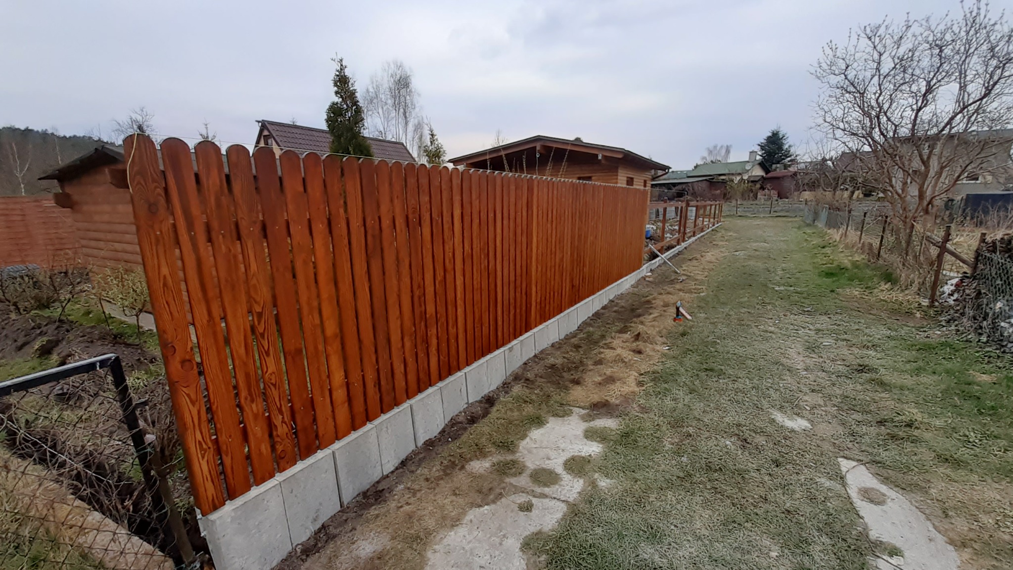 Nowo postawione drewniane ogrodzenie z palików, osadzone na betonowej podmurówce, widoczne na tle zabudowy jednorodzinnej w pochmurny dzień.