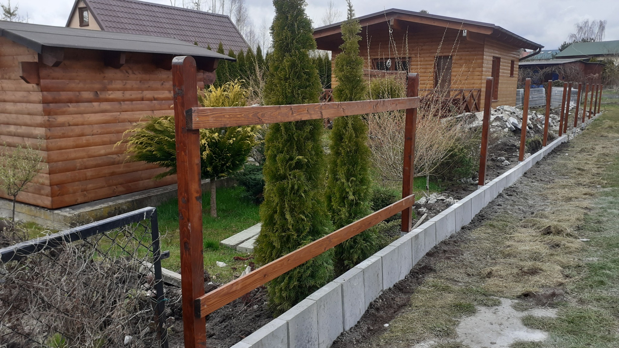 Wznoszenie drewnianego ogrodzenia z betonową podmurówką na działce z domkami letniskowymi i tujami.