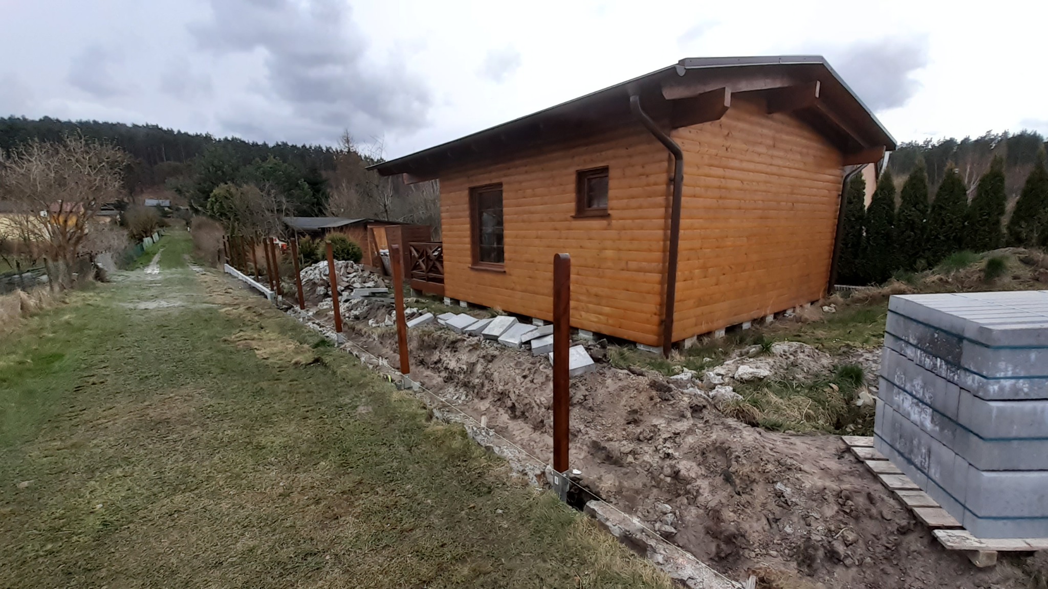 Montaż ogrodzenia: drewniane słupki ogrodzeniowe ustawione w wykopie wzdłuż drewnianego domku letniskowego, widoczne płyty chodnikowe i stos kostki brukowej, w tle las