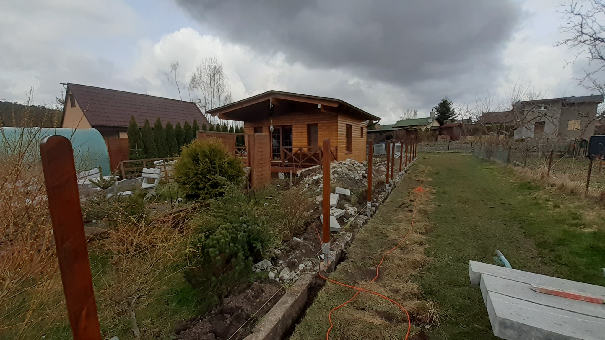 Wykop pod fundament ogrodzenia z drewnianymi słupkami w trakcie realizacji, widoczny sznurek wyznaczający linię, w tle drewniany domek letniskowy i pochmurne niebo.