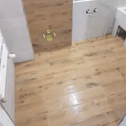 Ułożone płytki podłogowe imitujące drewno w łazience, z widocznym fragmentem ściany z białymi płytkami imitującymi marmur oraz zabudową geberit z płytkami drewnopodobnymi i elementami instalacji.