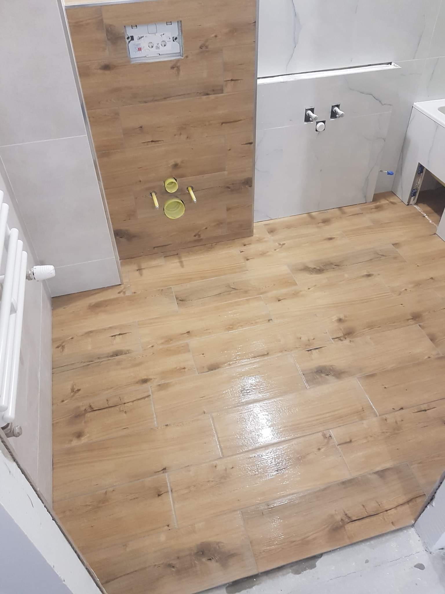 Ułożone płytki podłogowe imitujące drewno w łazience, z widocznym fragmentem ściany z białymi płytkami imitującymi marmur oraz zabudową geberit z płytkami drewnopodobnymi i elementami instalacji.