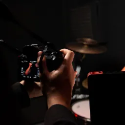 Kamerzysta filmuje perkusistę w studio, widoczny ekran aparatu z ustawieniami ekspozycji i ostrości