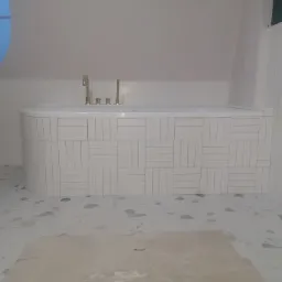 Biała wanna obłożona prostokątnymi płytkami w jodełkę, złota armatura, okrągłe okno w skosie dachu, podłoga z lastryko, widoczny fragment niezakończonej podłogi.