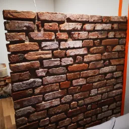 Ściana działowa z cegły klinkierowej w salonie, widoczne fragmenty kanapy i pomarańczowego paska na ścianie, sufit z kratką wentylacyjną.