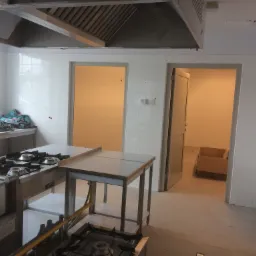 Wnętrze kuchni przemysłowej w trakcie prac wykończeniowych, widoczne płytki na ścianach, stalowe meble robocze, kuchenka gazowa i system wentylacyjny pod sufitem.