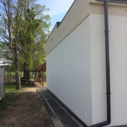 Elewacja budynku z jasnym tynkiem i ciemną rynną, widoczna kostka brukowa i zieleń w tle.