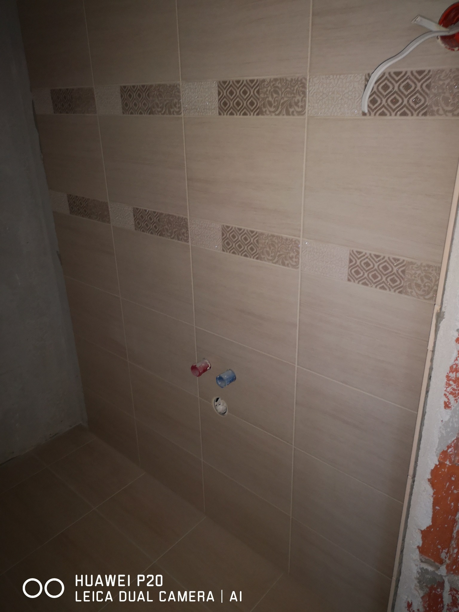 Ściana i podłoga łazienki wyłożone beżowymi płytkami z dekoracyjnym pasem, widoczne wystające rury instalacji wodnej i fragment instalacji elektrycznej w trakcie remontu.