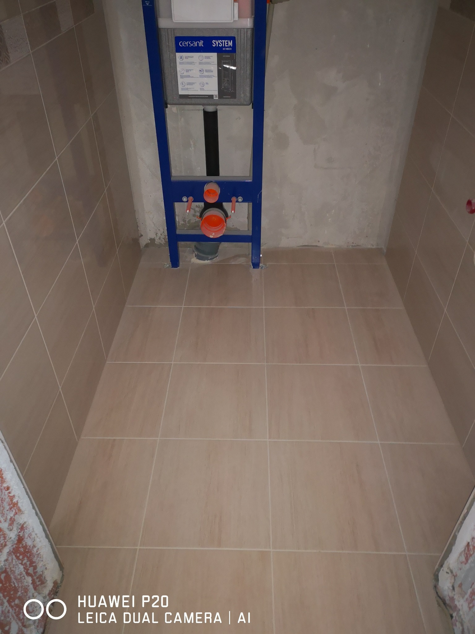 Beżowe płytki podłogowe i częściowo wykończone ściany w małej łazience, z widoczną niebieską konstrukcją podtynkową WC Cersanit System i instalacją hydrauliczną.