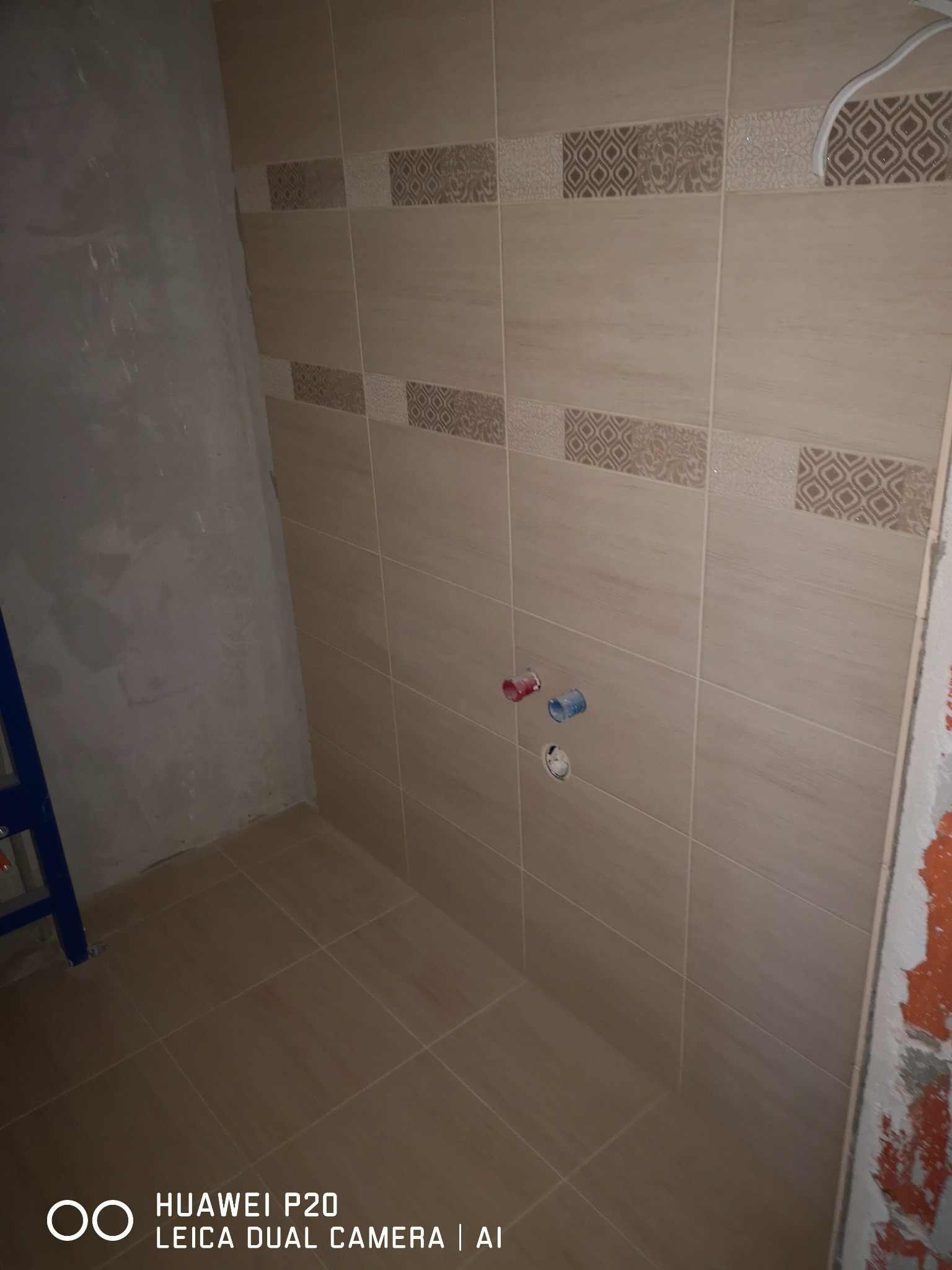 Ściana łazienki w trakcie układania płytek, widoczne przygotowane przyłącza wodne i fragment surowej ściany z resztkami farby.