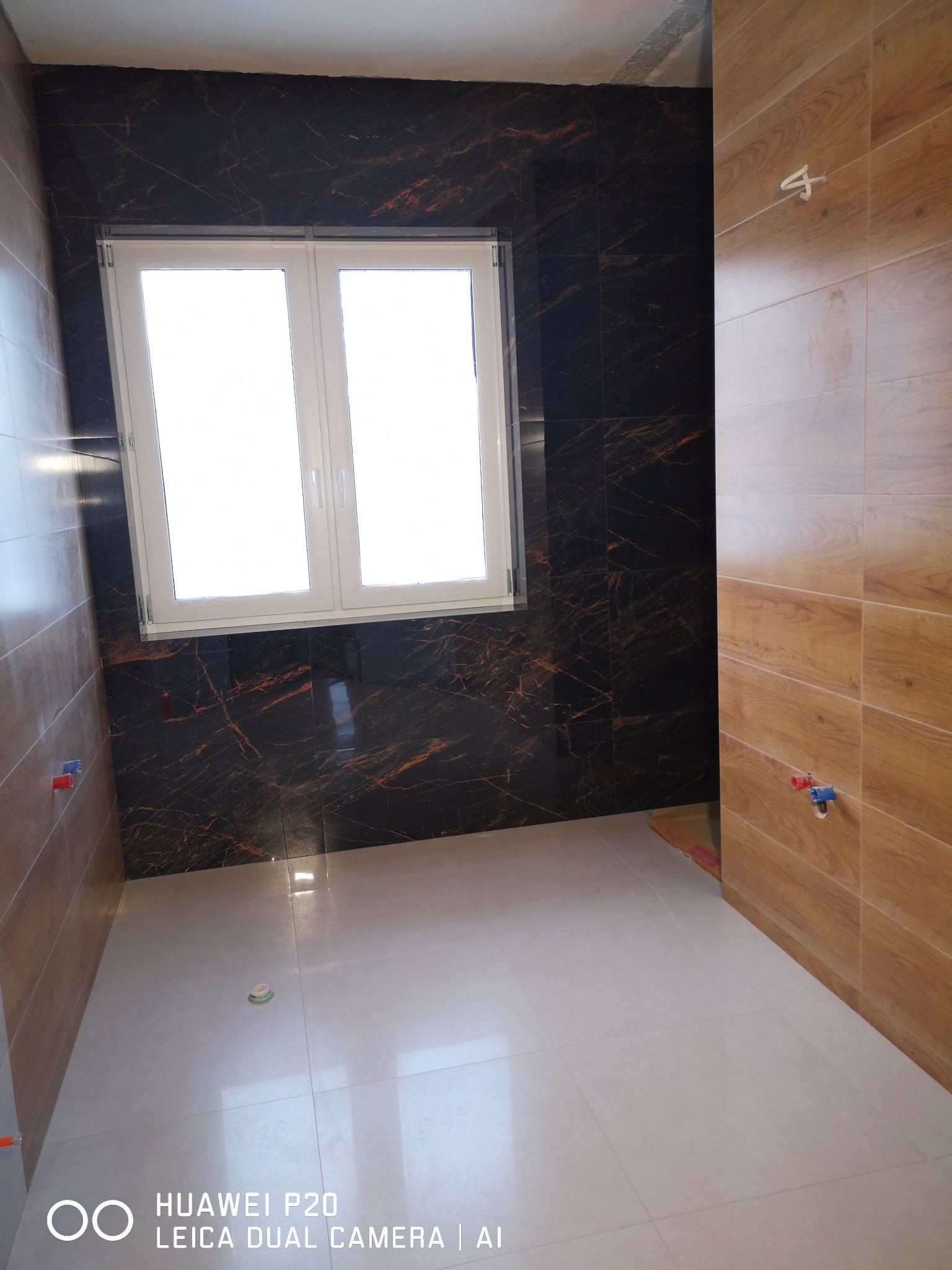 Wykończona łazienka z czarnymi płytkami imitującymi marmur na ścianie z oknem, drewnopodobnymi płytkami na sąsiedniej ścianie i jasnymi płytkami podłogowymi. Widoczne podejścia wodne.