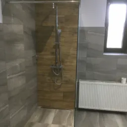 Nowoczesna kabina prysznicowa walk-in z deszczownicą, wykończona płytkami imitującymi drewno i szarym marmurem, z grzejnikiem panelowym i oknem w tle.