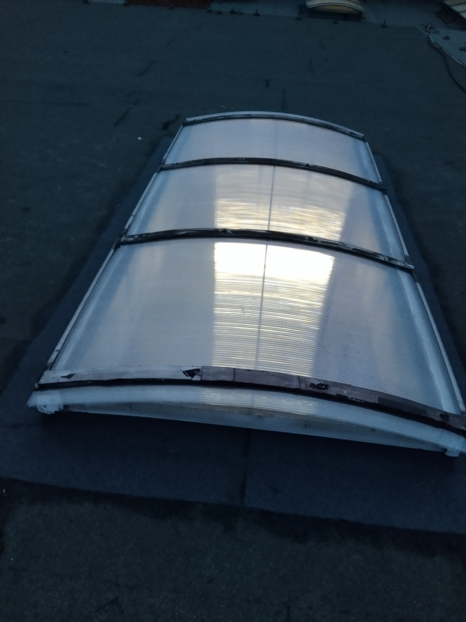 Półprzezroczyste okno dachowe z widocznymi uszkodzeniami ramy, osadzone na płaskim dachu pokrytym ciemną papą, odbijające światło słoneczne.