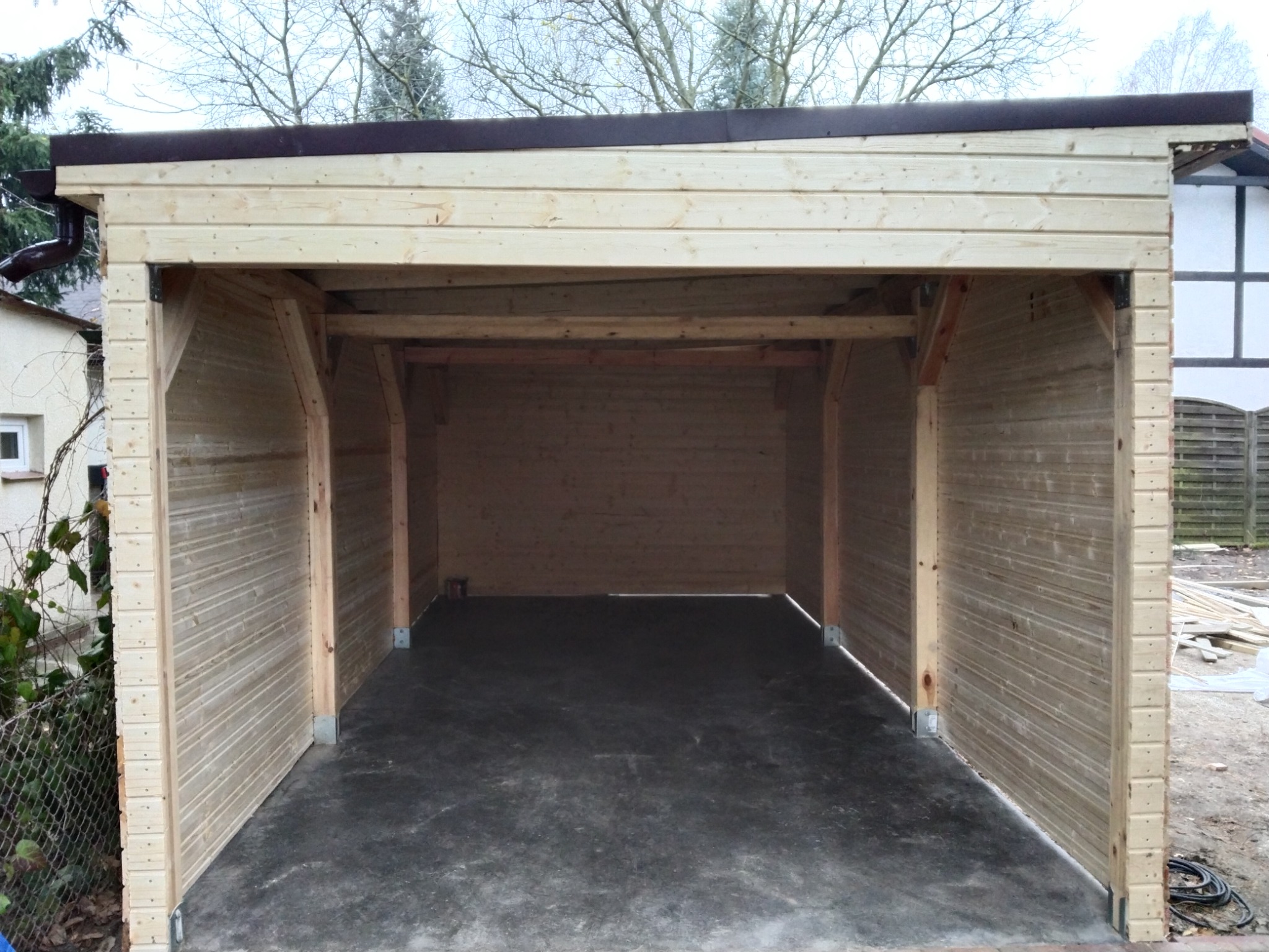 Niedokończona konstrukcja drewnianej wiaty garażowej z widoczną konstrukcją dachu i ścian z desek, surowy beton na podłodze, widok z zewnątrz.