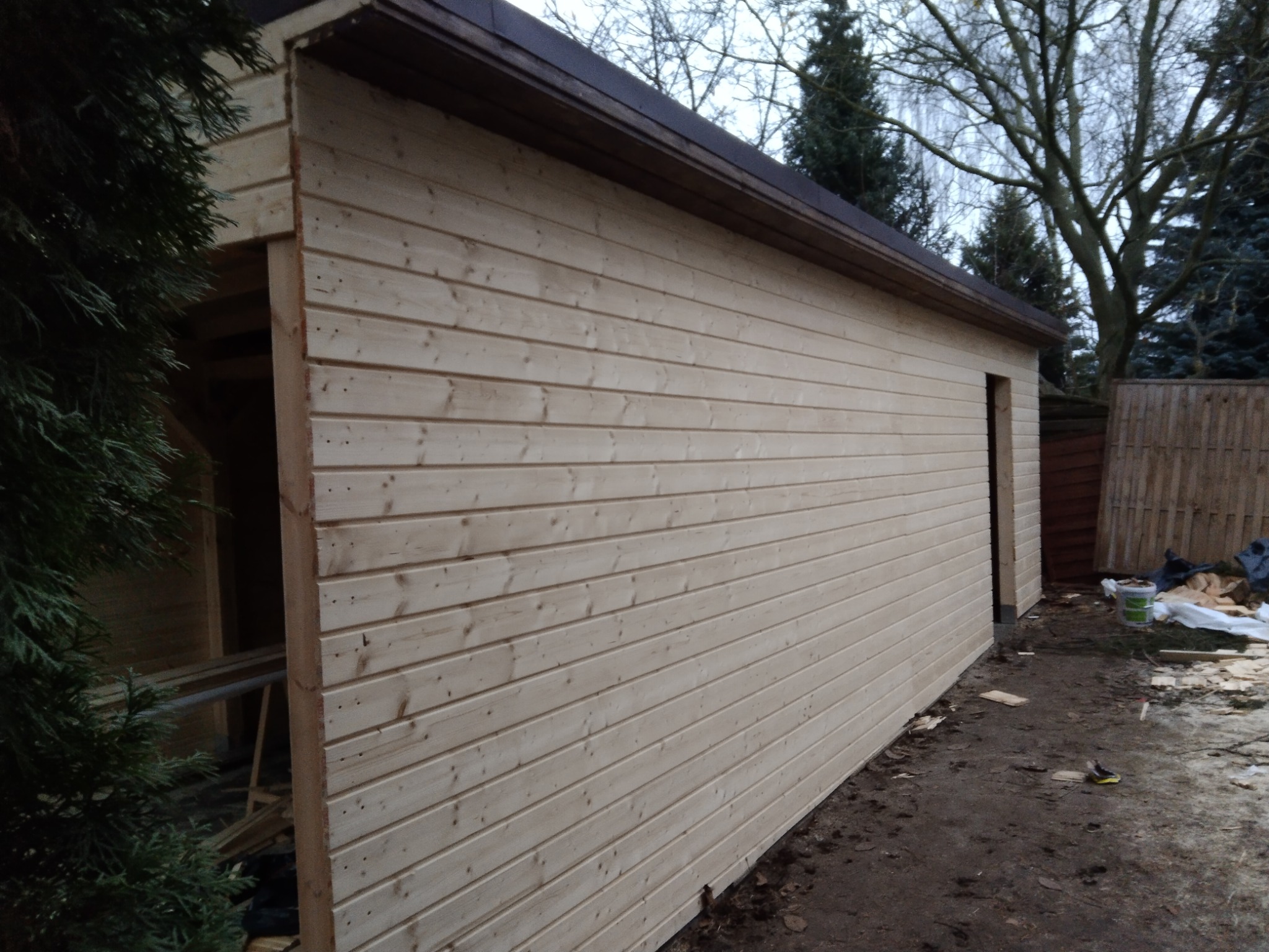 Nowo wybudowany, jasny drewniany budynek gospodarczy z brązowym dachem, widoczny z boku, z elementami wykończeniowymi i pozostawionymi narzędziami na ziemi.