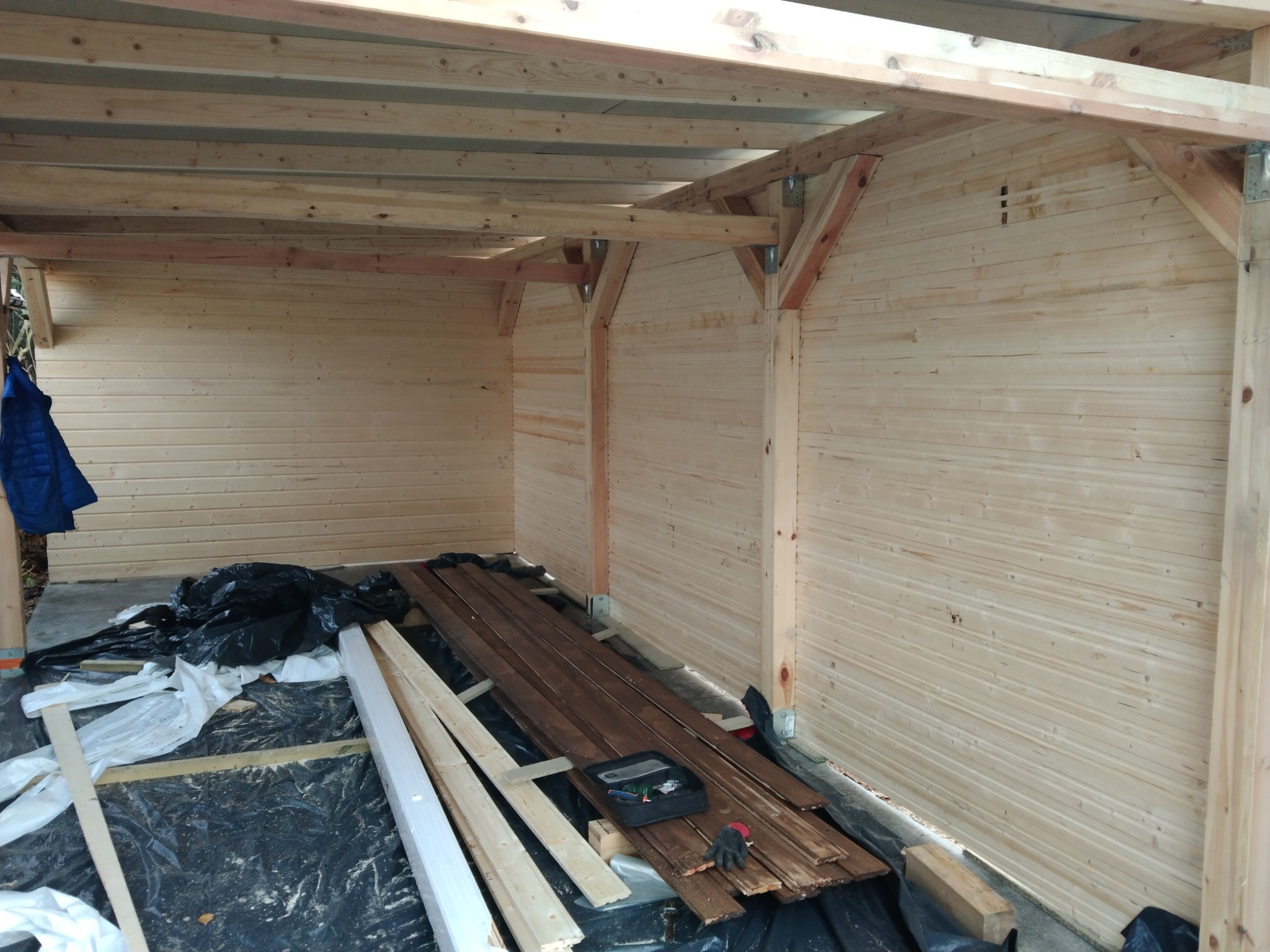 Wnętrze drewnianej konstrukcji szkieletowej w trakcie budowy, widoczne deski ścienne i stropowe, narzędzia i materiały budowlane na podłodze zabezpieczonej folią, niebieska kurtka wisząca z boku.