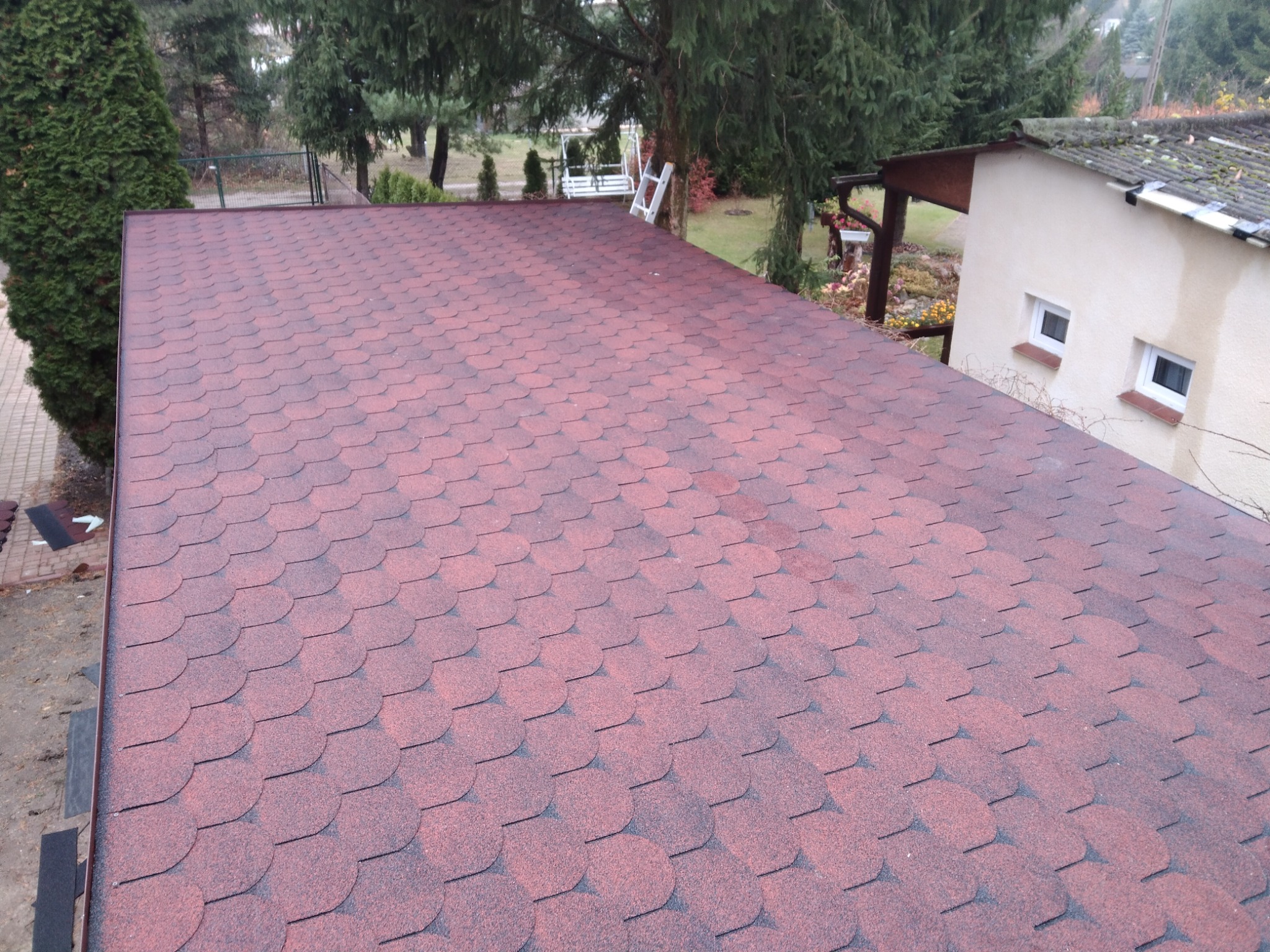 Nowy dach pokryty czerwoną dachówką bitumiczną, widok z góry na tle zieleni ogrodowej i fragmentu budynku.