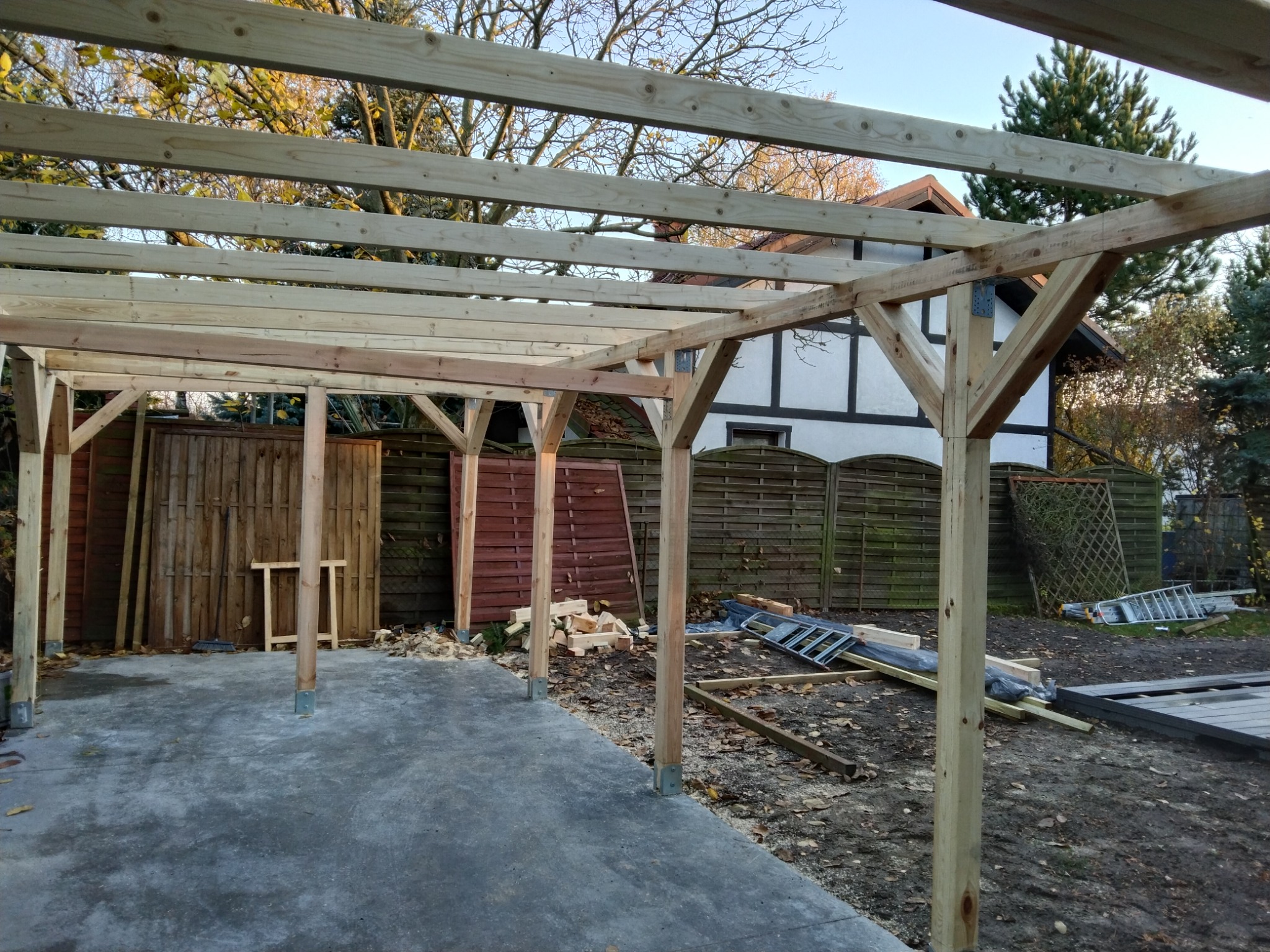 Konstrukcja drewnianego zadaszenia w trakcie budowy, widoczne słupy i belki, w tle dom z elementami muru pruskiego i ogrodzenie.