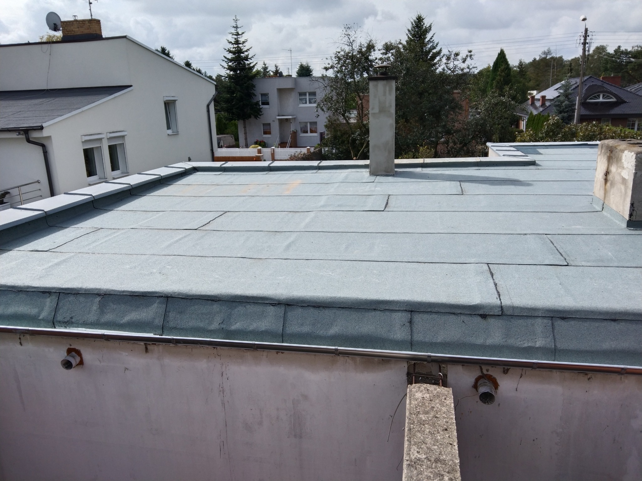 Płaski dach pokryty papą w kolorze szaro-zielonym, widoczny fragment komina i rynna z odpływem, w tle zabudowa jednorodzinna pod pochmurnym niebem.