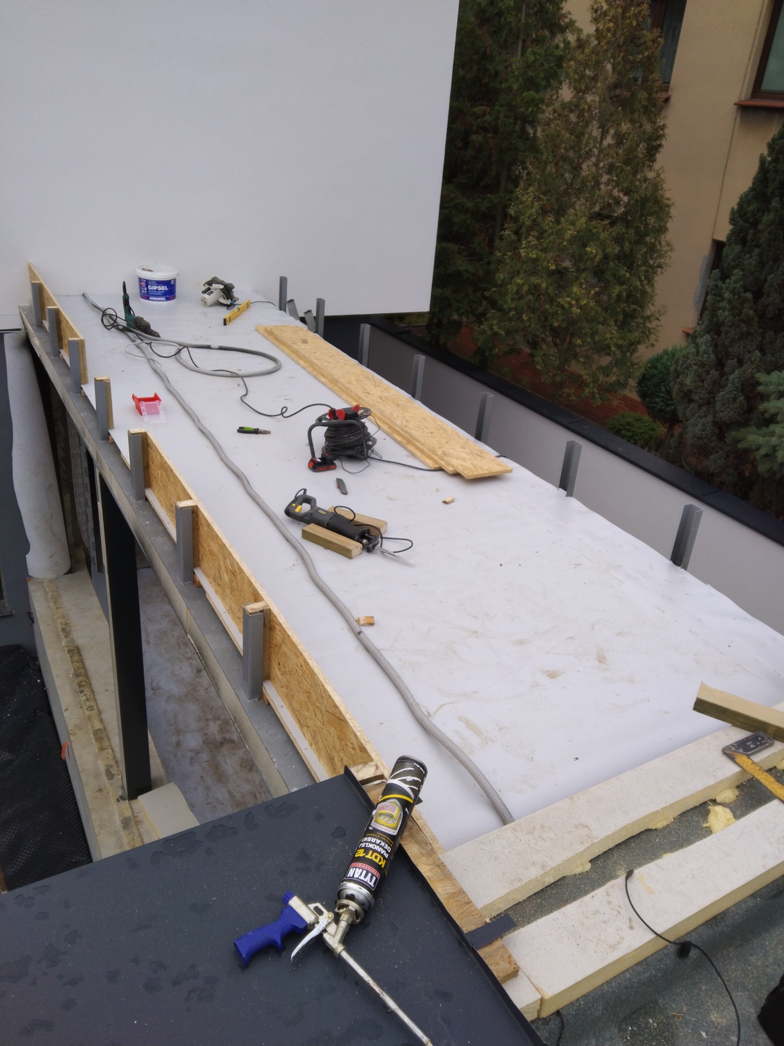 Montaż izolacji na dachu płaskim z użyciem pianki montażowej, widoczna konstrukcja z drewna i metalu, narzędzia pracy oraz częściowo ułożona warstwa hydroizolacyjna.