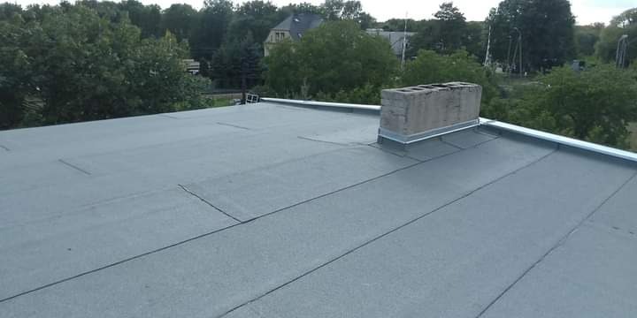 Płaski dach pokryty papą termozgrzewalną z widocznym kominem obłożonym blachą i obróbką blacharską na krawędzi dachu, w tle zieleń drzew.