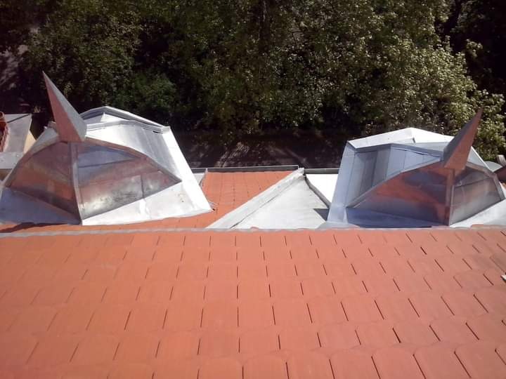 Czerwony dach z dachówki ceramicznej z dwoma metalowymi lukarnami w kształcie piramidy, widok z góry, w tle zieleń drzew.