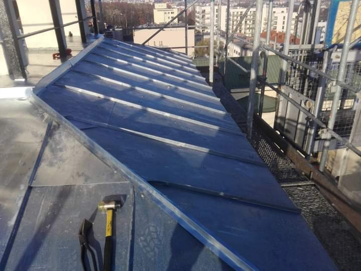 Szary dach z blachy na budynku, widoczne narzędzia dekarskie - młotek i szczypce, rusztowanie w tle, perspektywa z góry.