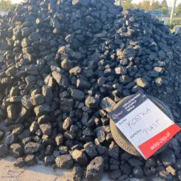 Góra czarnego węgla kamiennego na placu, z kartką informacyjną o nazwie 'Kostka Piast' i logo PGG, leżącą na oponie.