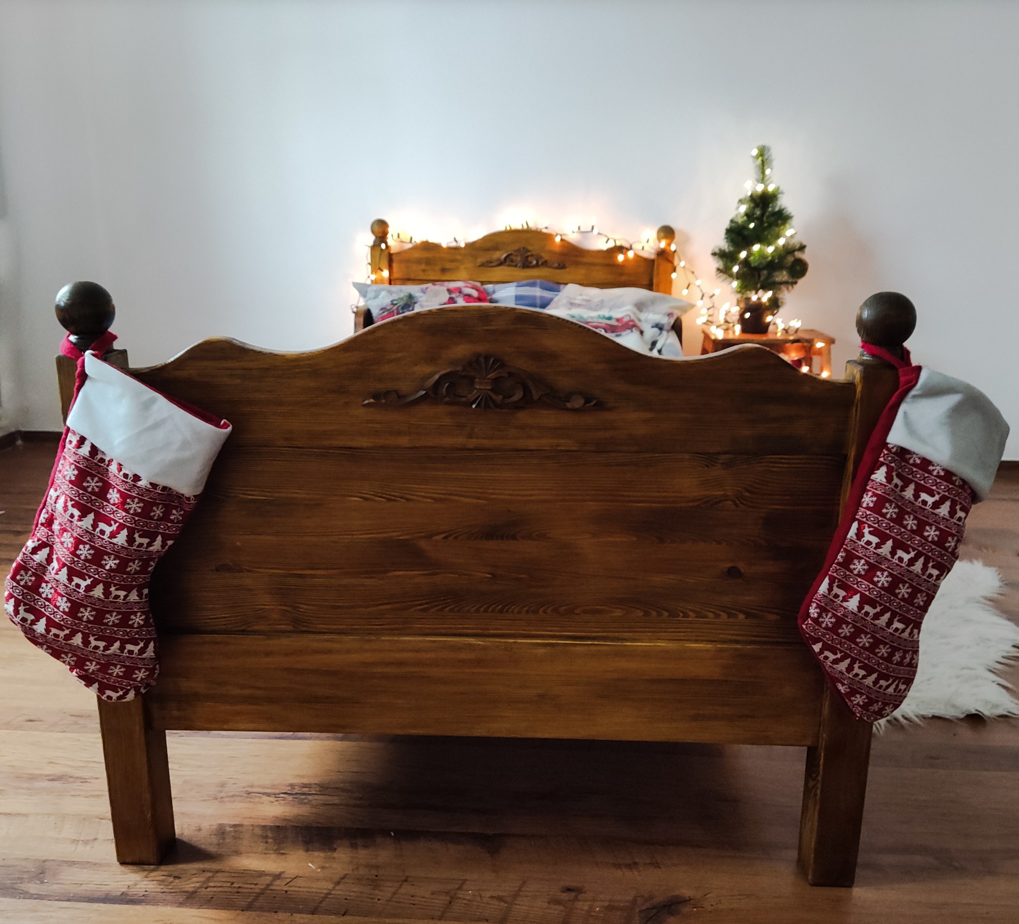 Drewniane łóżko z ręcznie rzeźbionym wezgłowiem, udekorowane świątecznymi skarpetami i lampkami, z małą choinką w tle.