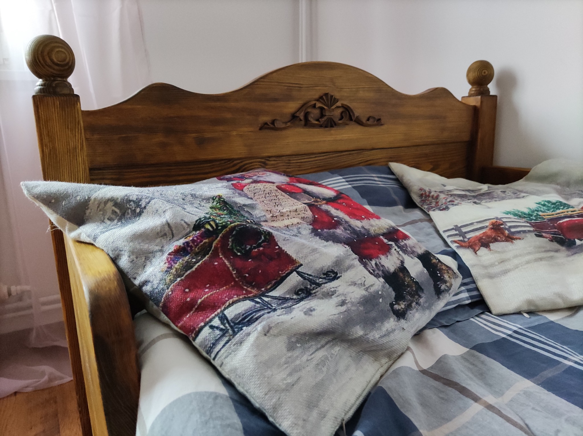 Fragment łóżka z drewnianym wezgłowiem, na którym leżą poduszki z motywami świątecznymi, w tym wizerunek Świętego Mikołaja i sań.