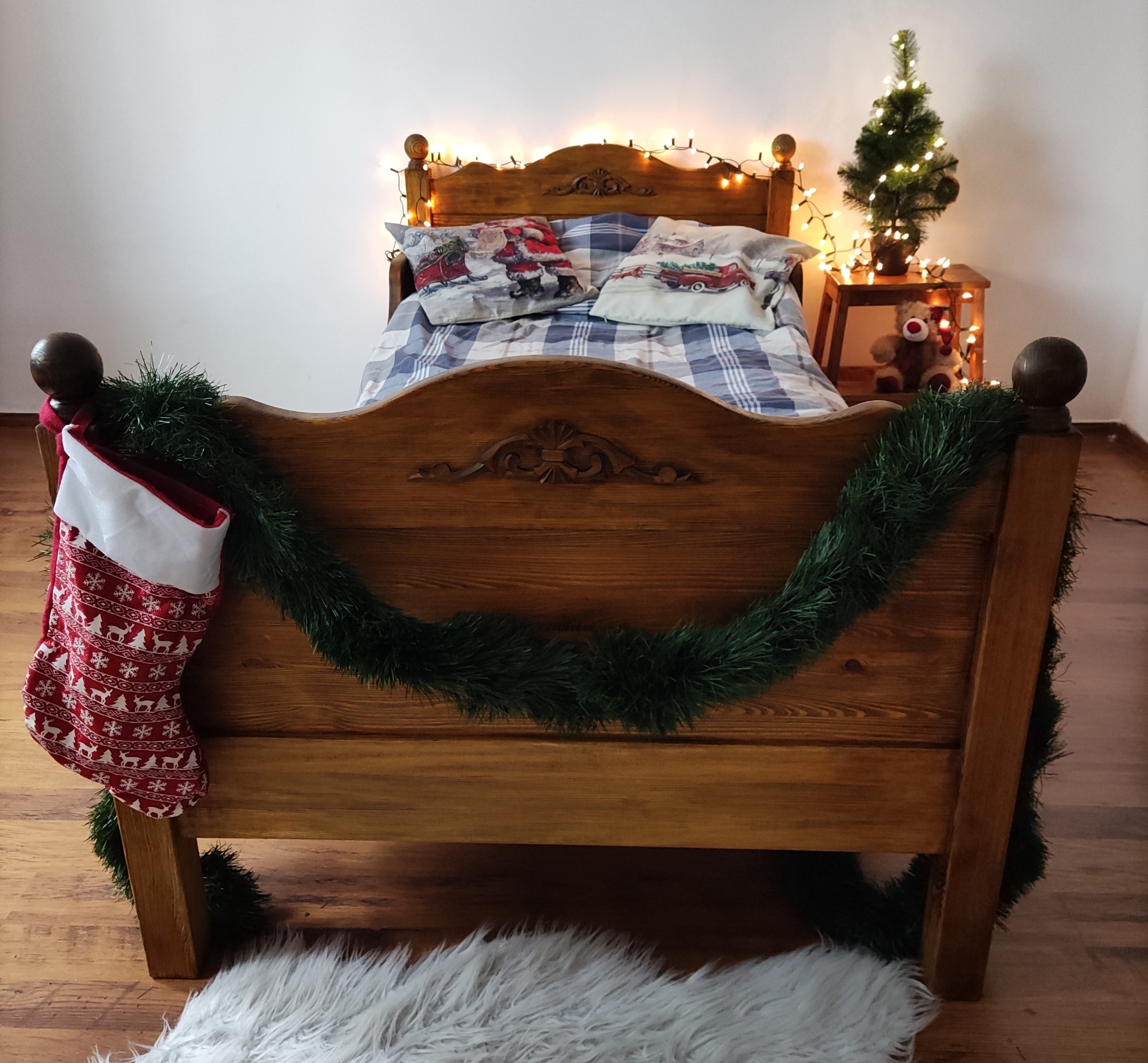 Drewniane łóżko z dekoracjami świątecznymi: skarpeta z reniferami, girlanda z igliwia i mała choinka z lampkami.