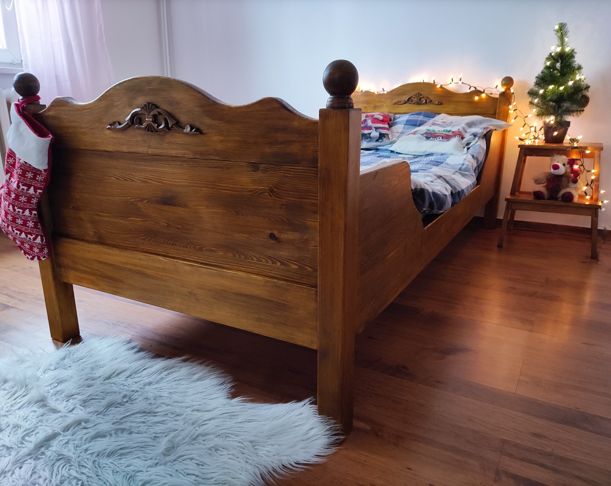 Drewniane łóżko z ozdobnym wezgłowiem i świąteczną skarpetą, na podłodze puszysty dywanik, w tle mała choinka z lampkami i pluszowy renifer na krześle.