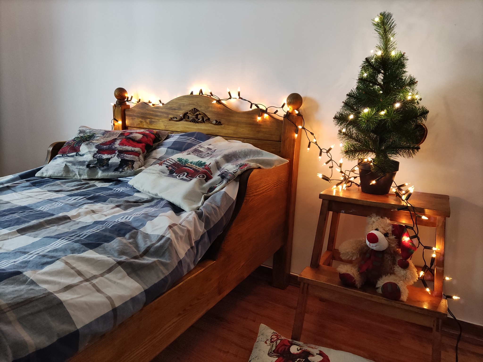 Drewniane łóżko z pościelą w kratę i poduszkami ze świątecznym motywem, oświetlone lampkami, obok drewniany stołek z małą choinką i pluszowym reniferem, również oświetlone lampkami.