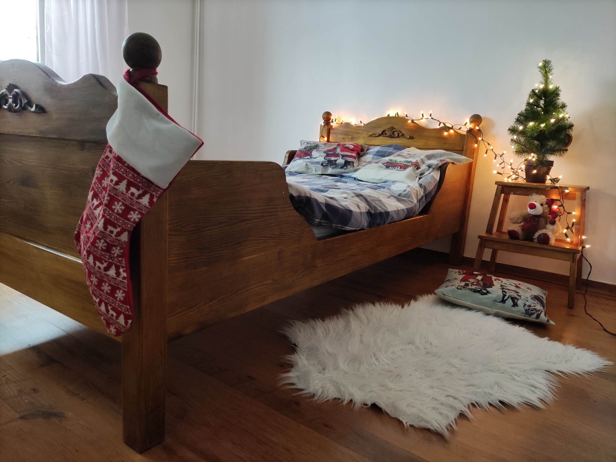 Drewniane łóżko dziecięce z świątecznymi dekoracjami: skarpeta na prezenty, poduszki z motywem świątecznym, choinka z lampkami na stoliku obok.