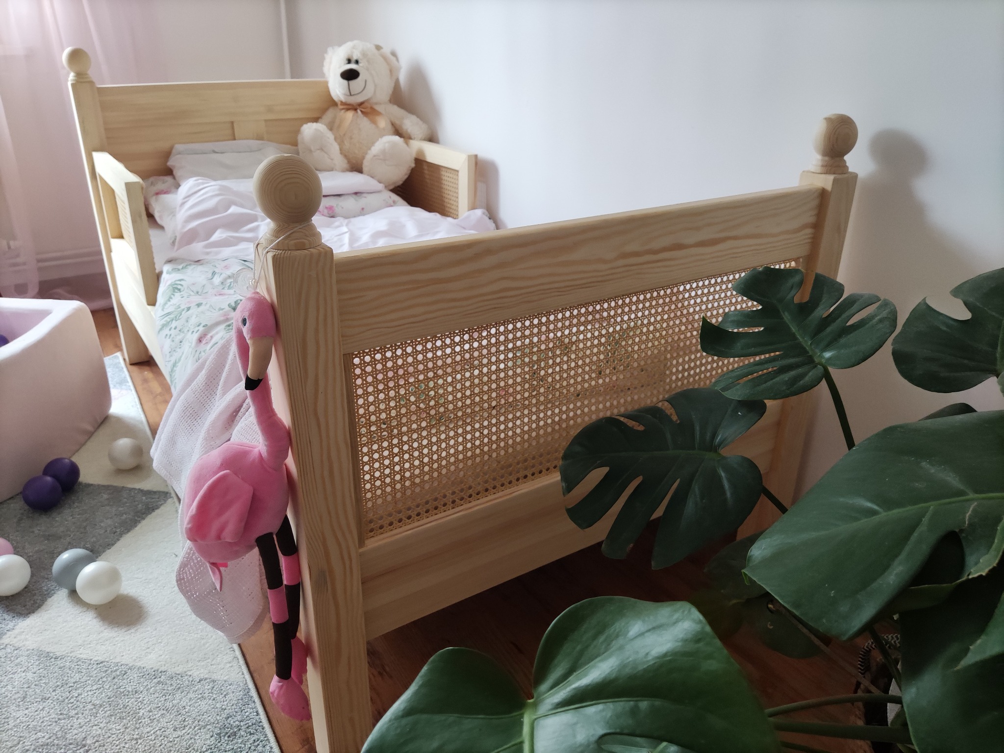 Drewniane łóżeczko dziecięce z rattanowym zagłówkiem, pluszowy miś na poduszce, różowy flaming-zabawka zwisający z boku, obok doniczka z monsterą.