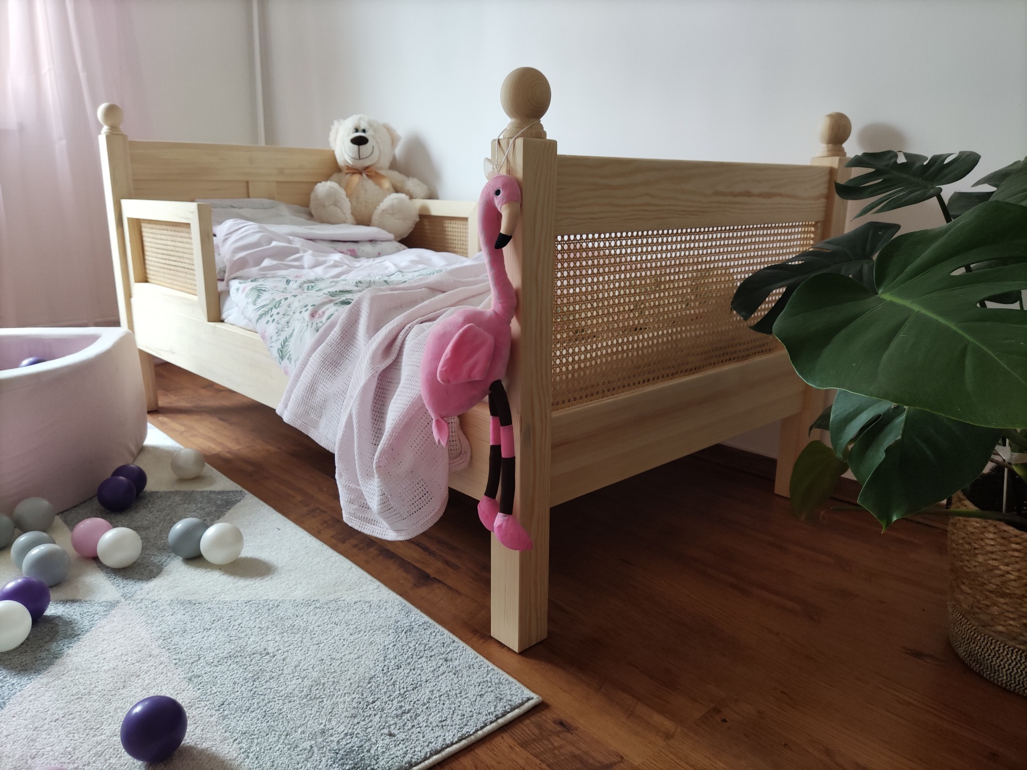 Drewniane łóżko dziecięce z rattanowym zagłówkiem, ozdobione pluszowym flamingiem i misiem, w pokoju z drewnianą podłogą, dywanem w geometryczne wzory i rośliną doniczkową.