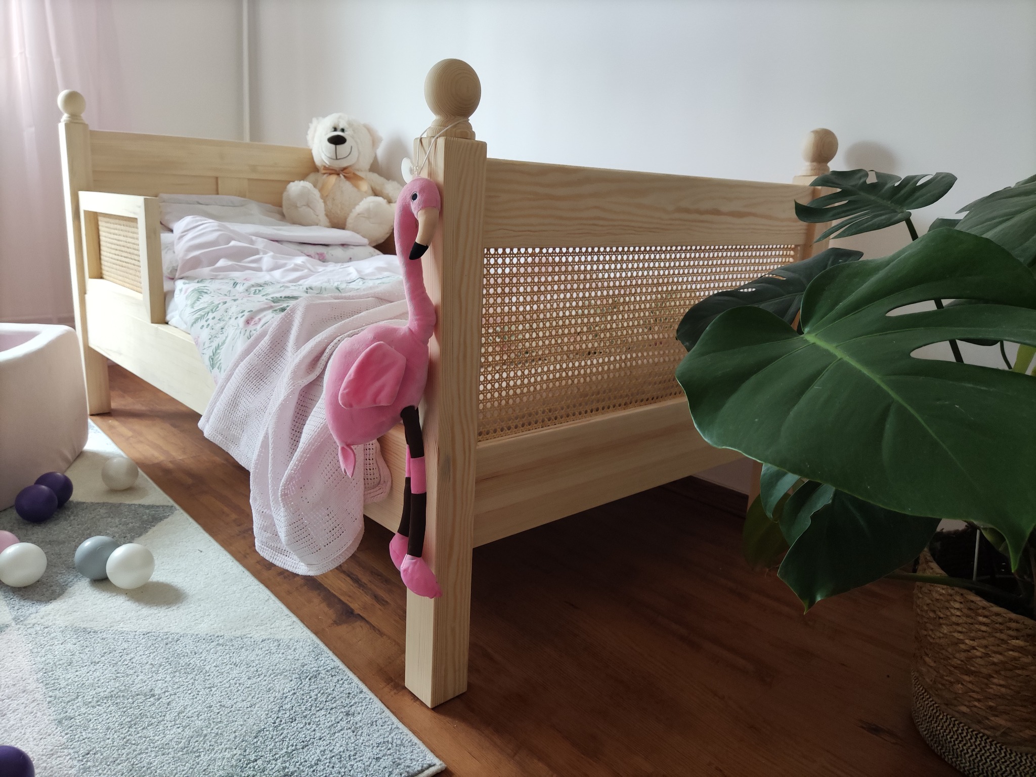 Drewniane łóżko dziecięce z rattanowym zagłówkiem, ozdobione pluszowym flamingiem i misiem, na podłodze dywan z kulkami do zabawy, obok doniczka z monsterą.