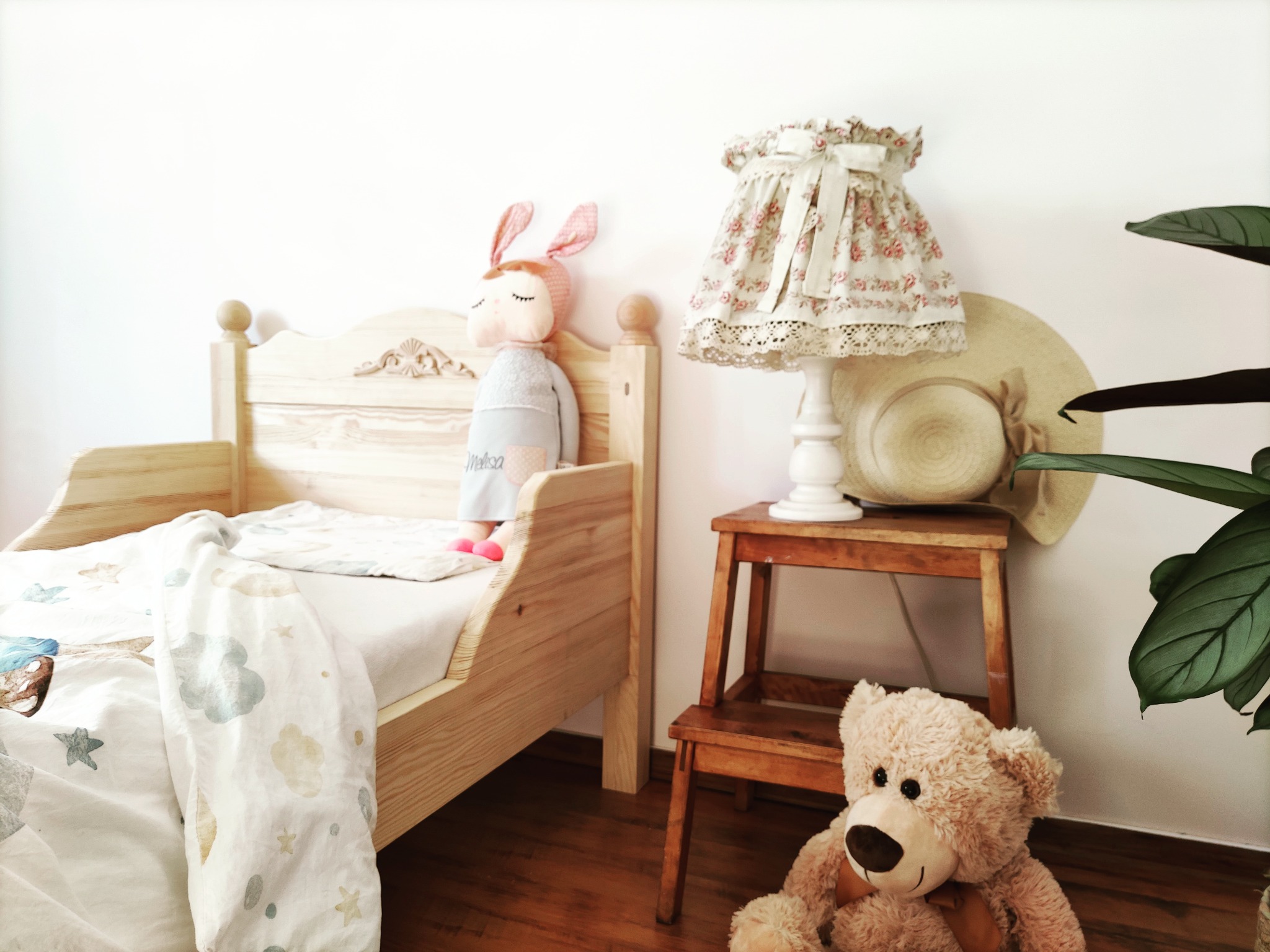 Drewniane łóżko dziecięce z pluszowym królikiem, lampką nocną z abażurem w kwiaty i pluszowym misiem na podłodze. Na ścianie wisi słomkowy kapelusz.