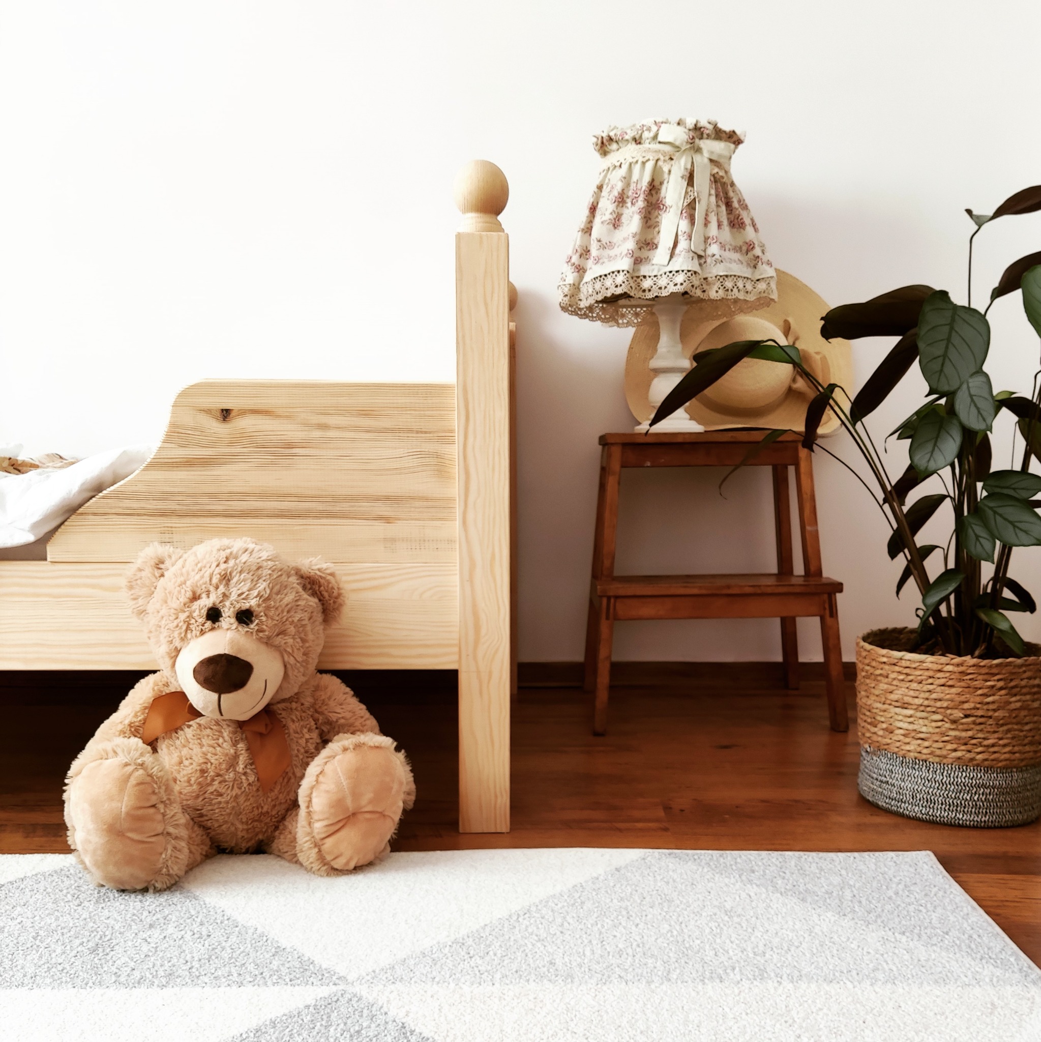 Fragment pokoju dziecięcego z drewnianym łóżkiem, pluszowym misiem siedzącym na geometrycznym dywanie, lampką nocną w kwiaty na drewnianym stołku i rośliną w plecionym koszu.
