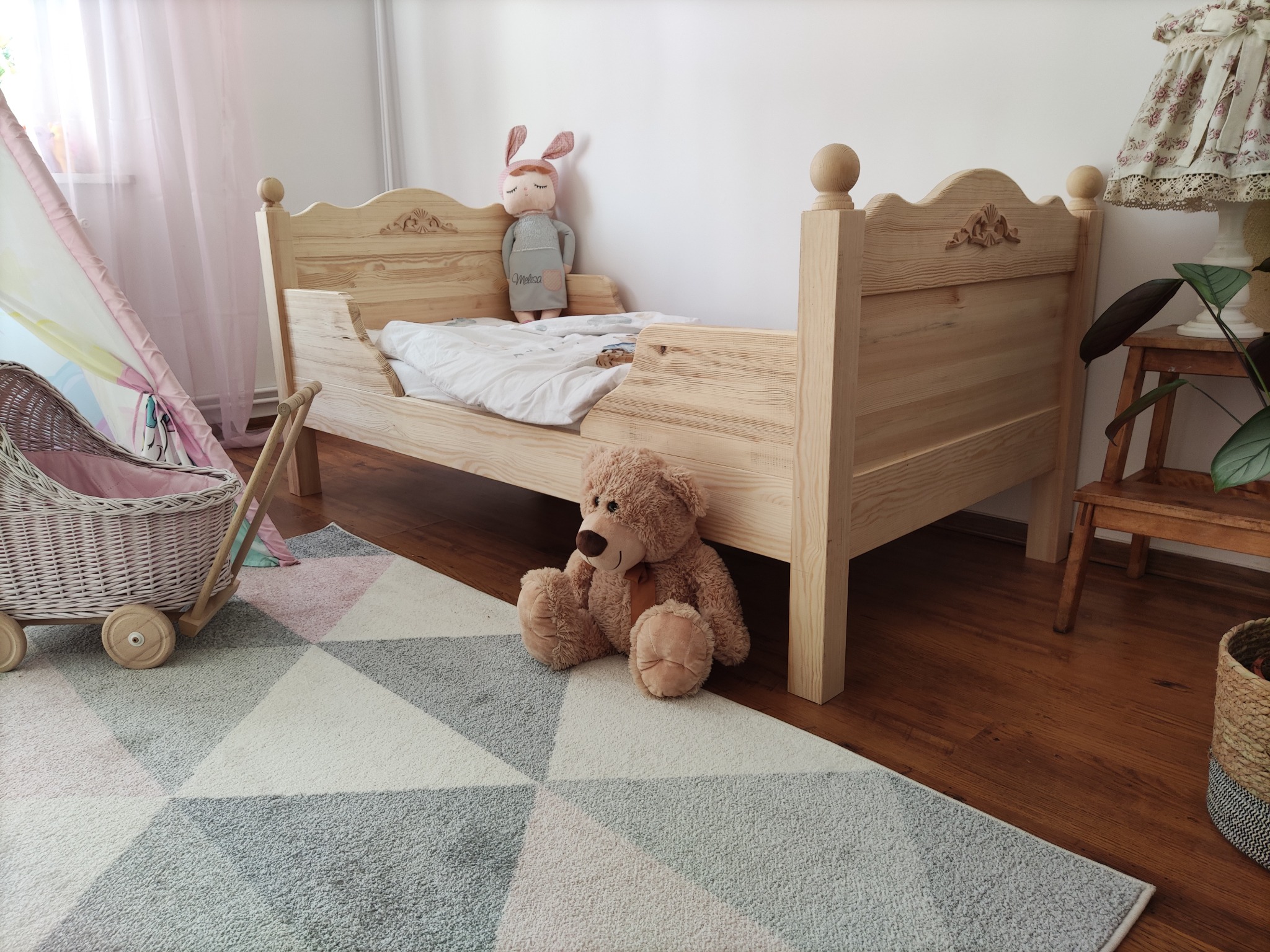 Drewniane łóżeczko dziecięce z rzeźbionym wezgłowiem, pluszowym misiem i lalką w pokoju w Stalowej Woli, na podłodze wzorzysty dywan.