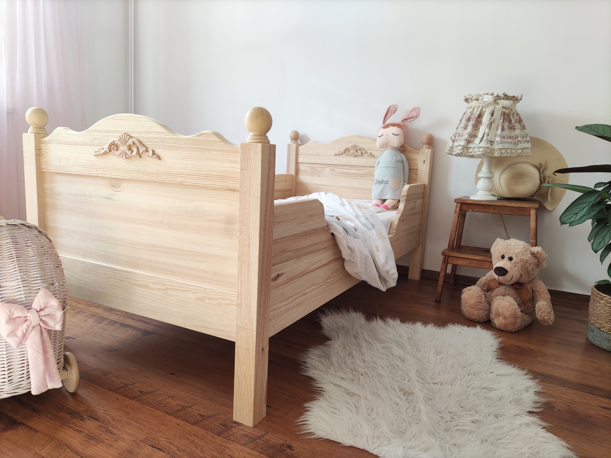 Drewniane łóżeczko dziecięce z dekoracyjnymi rzeźbieniami na wezgłowiu, ustawione w pokoju dziecięcym z pluszowym misiem, lampką nocną i dywanikiem z długim włosiem.